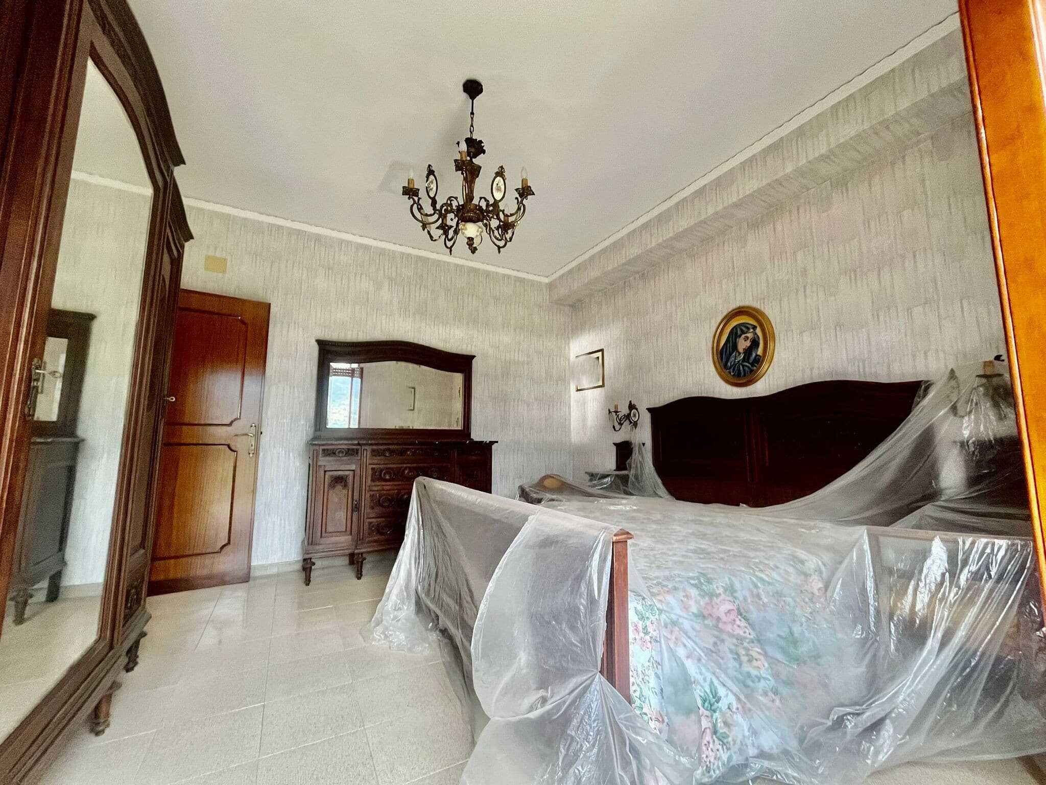 Letto matrimoniale in stile classico con tendaggi protettivi, mobili in legno scuro, specchio antico, pareti con carta da parati beige, lampadario elegante e quadro raffigurante una donna vestita di nero.