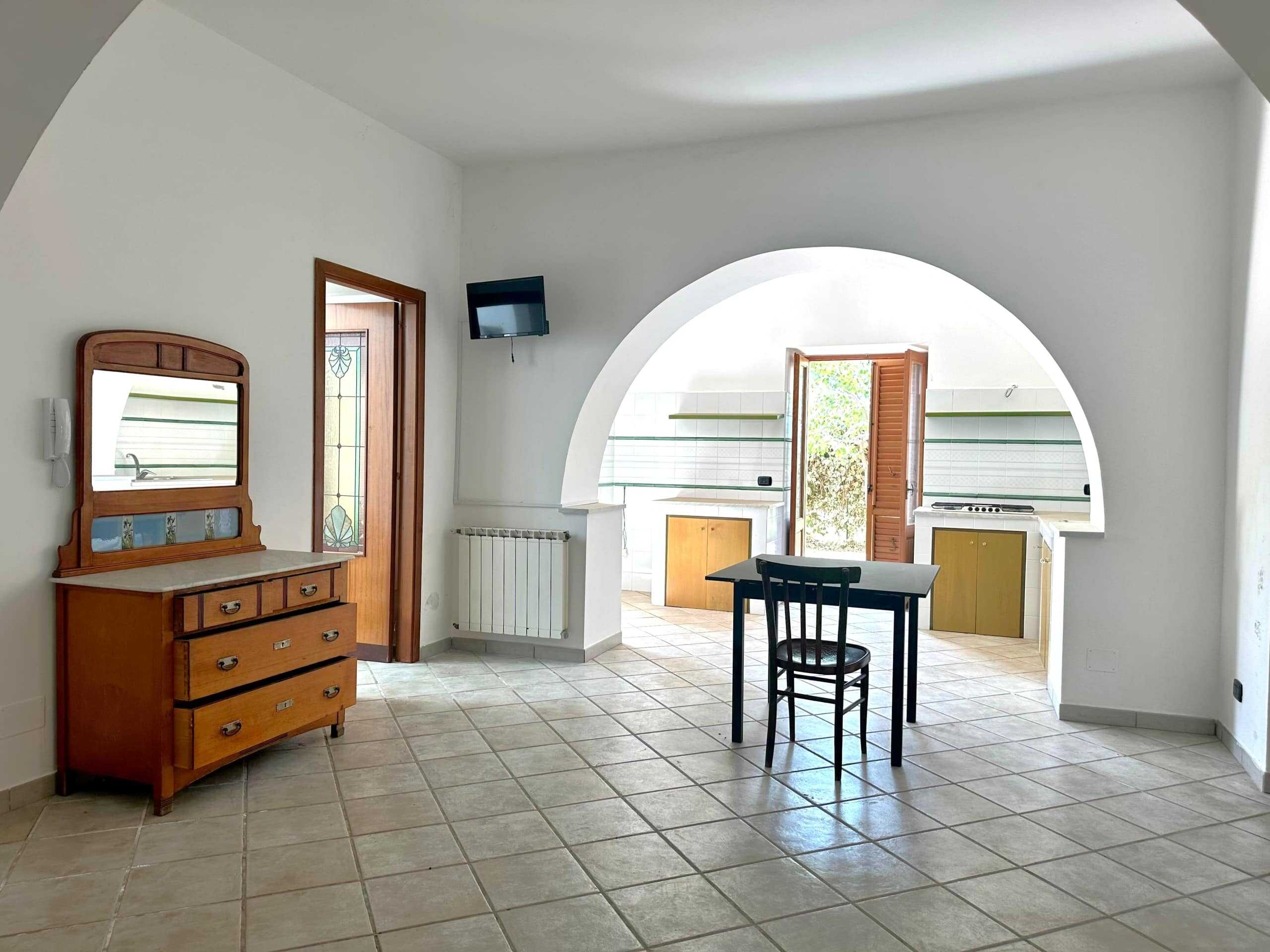 Arredo rustico soggiorno con accesso alla cucina in stile tradizionale, ampio spazio luminoso, pavimento in piastrelle beige, pareti bianche, grande arco e portafinestra che conducono all'esterno.