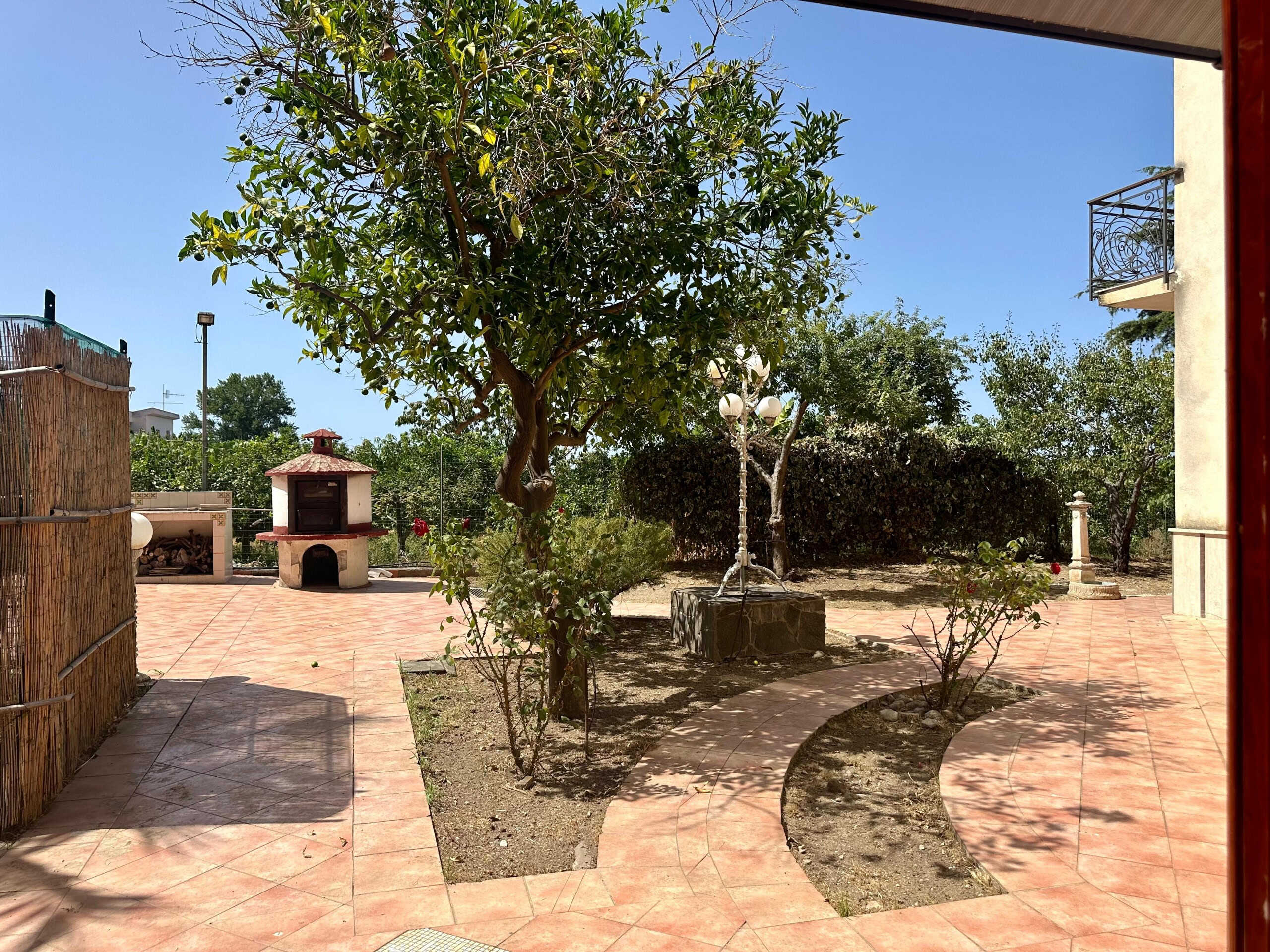 Cortile esterno con pavimentazione in ceramica, alberi verdi, illuminazione decorativa, barbecue in pietra e zone relax, ideale per vivere all'aperto e creare spazi conviviali.