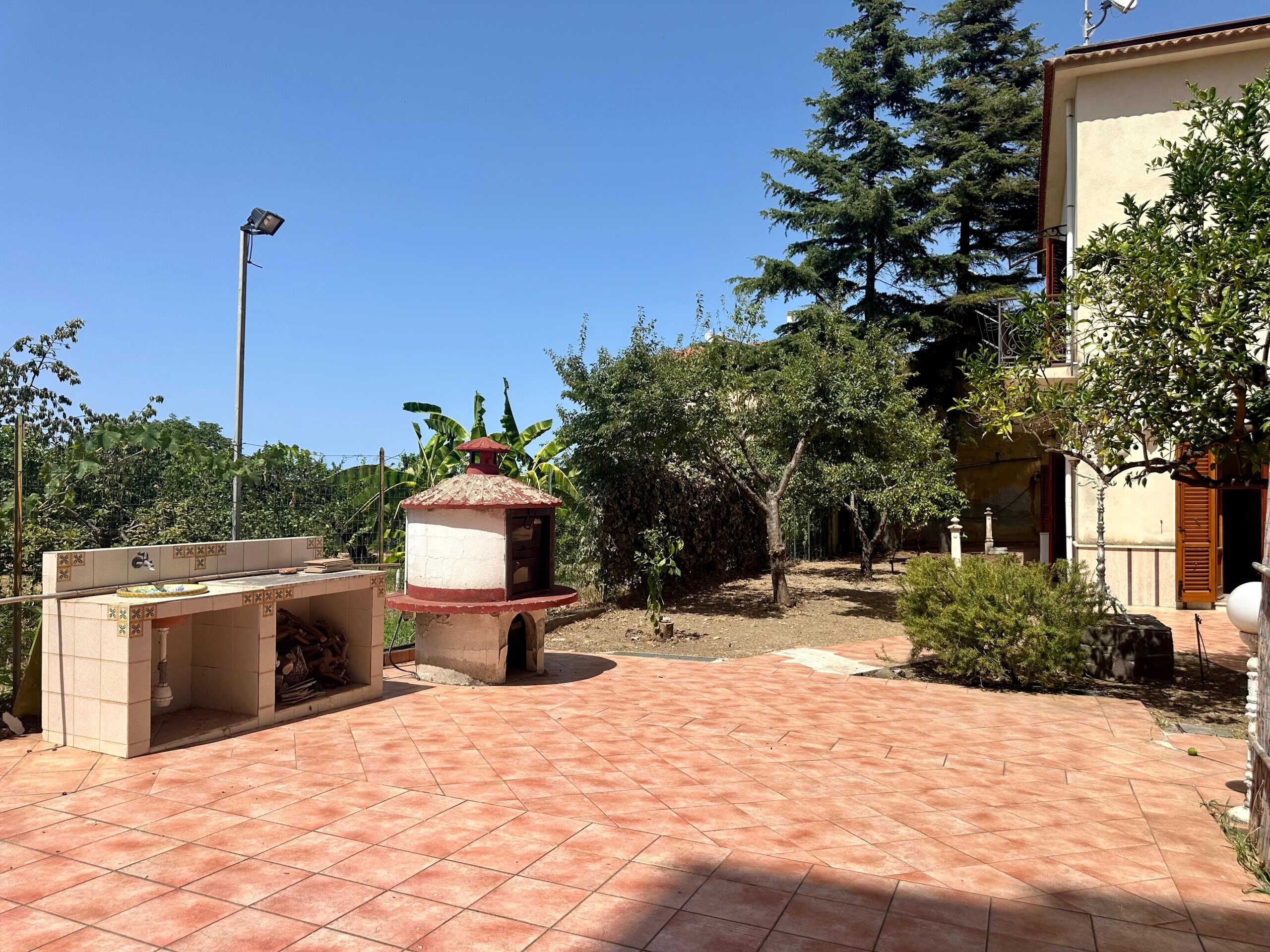 FOTO di cortile esterno con forno a legna, panchine, alberi, piante e ceramiche in mattonelle di terracotta, in un ambiente tranquillo e circondato da natura.
