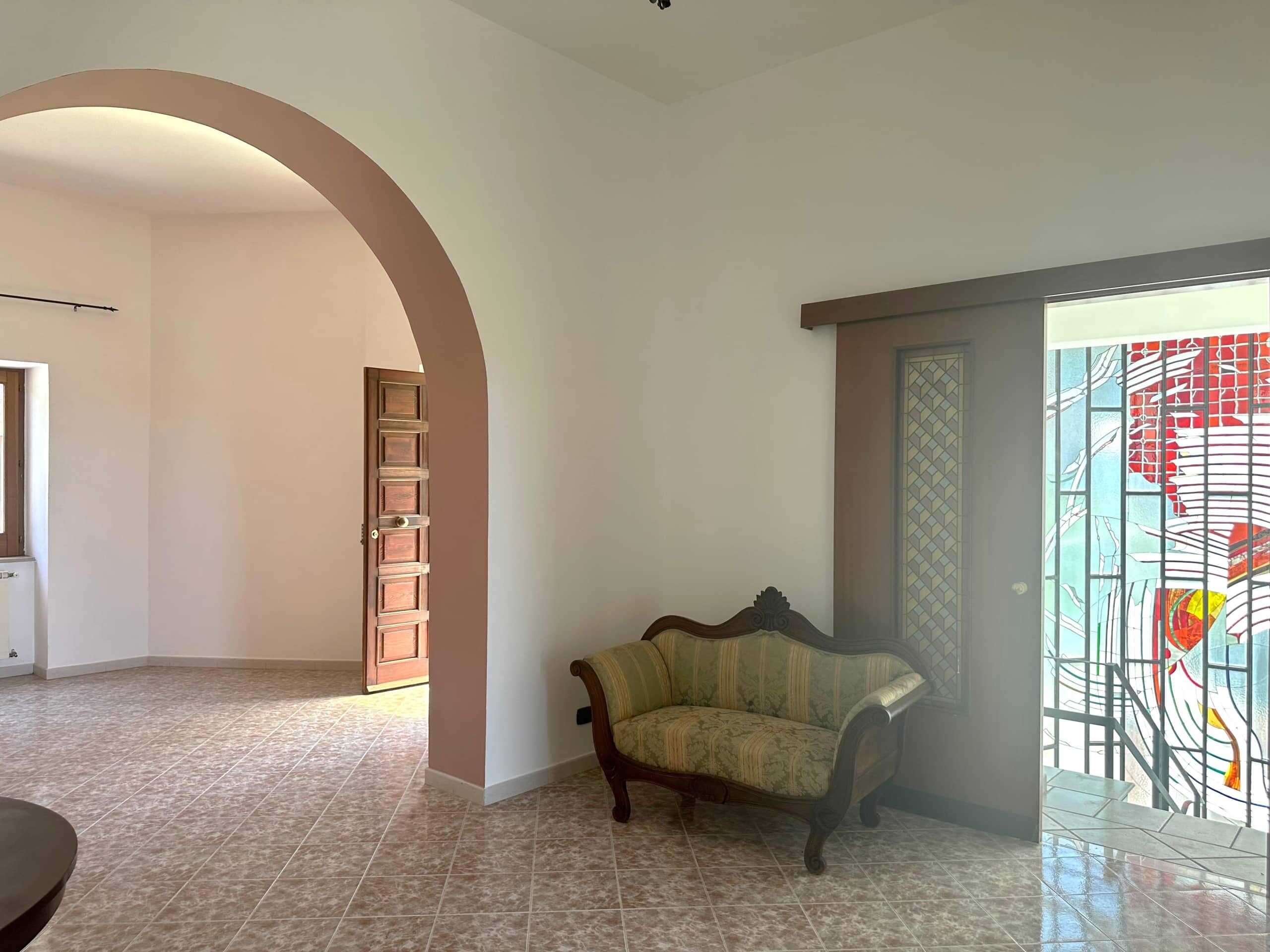 Arco in muratura che collega zona living e ingresso, interni luminosi e spaziosi con pavimento in ceramica, porta in legno e grande finestra con vetrata artistica nel soggiorno NextCasa.