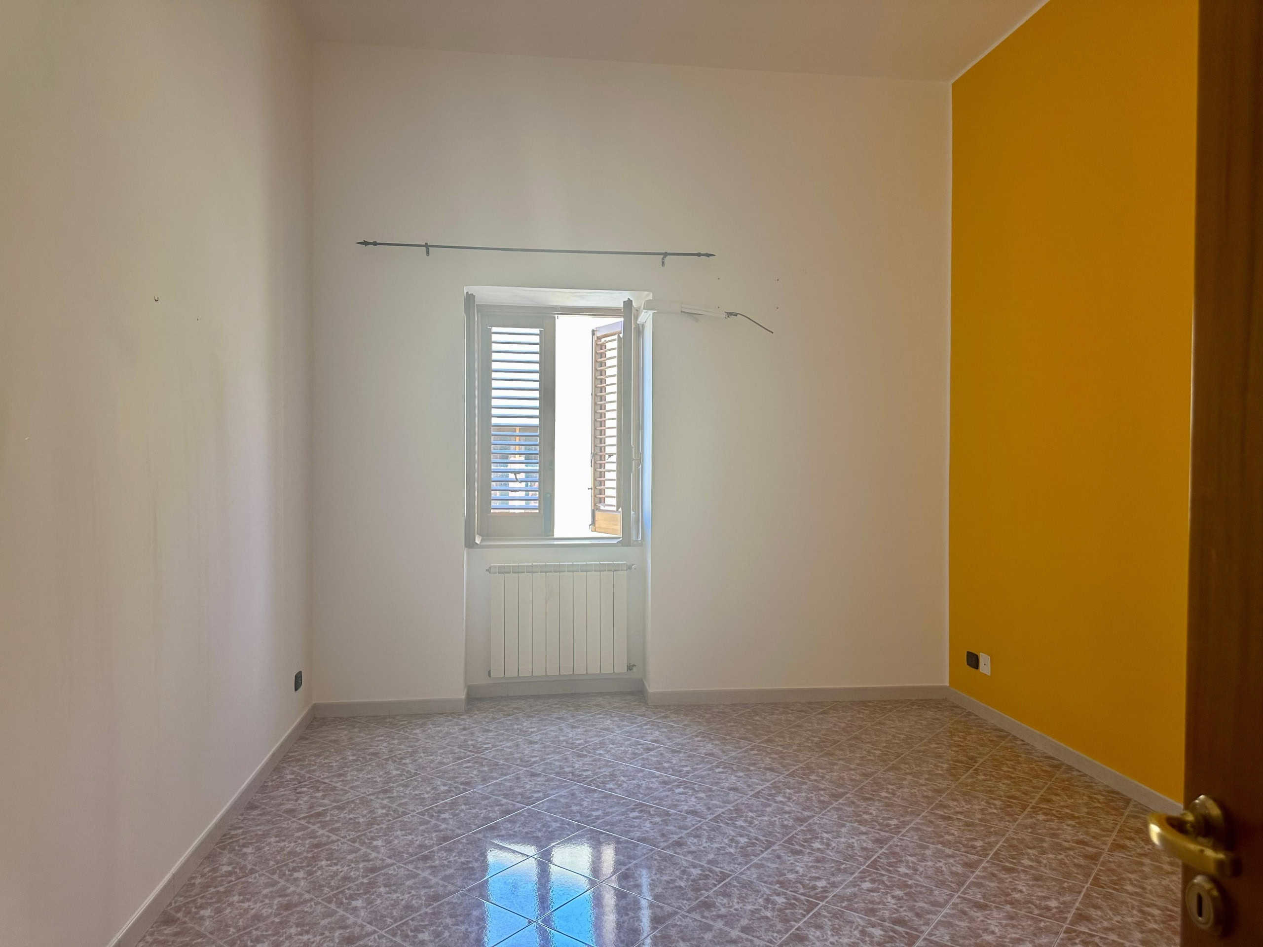 Finestra con persiane in una stanza vuota dall'aspetto luminoso, pareti bianche e una parete gialla, pavimento in gres e infissi moderni, pronta per arredare una casa moderna e accogliente.