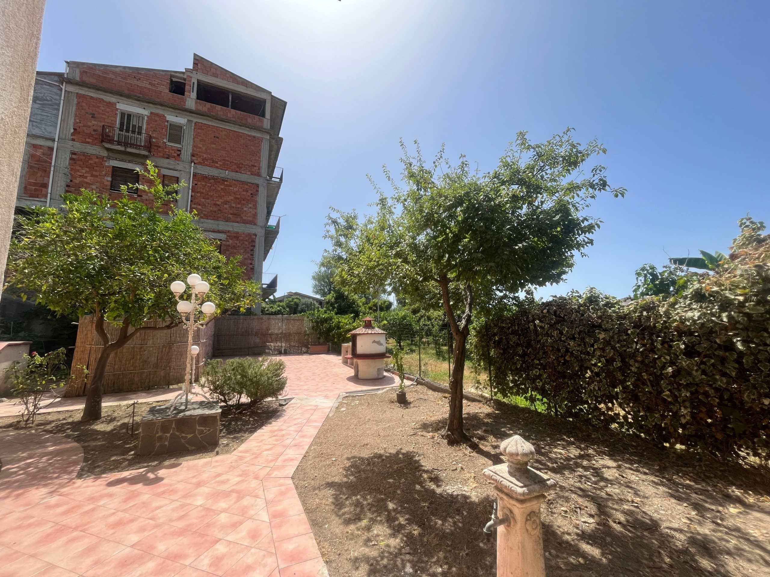 Veranda esterna di una casa con giardino, piante e area relax, con edifici di palazzi in costruzione in background, immersa in un ambiente tranquillo e naturale.