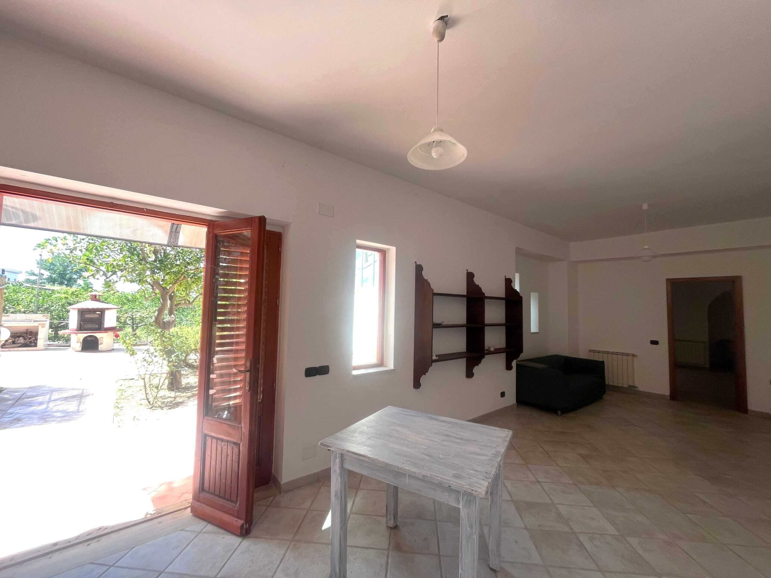 Ampia stanza luminosa con porta finestra che dà su un giardino, pareti bianche, soffitto con plafoniere, arredi minimalisti e pavimento in piastrelle.