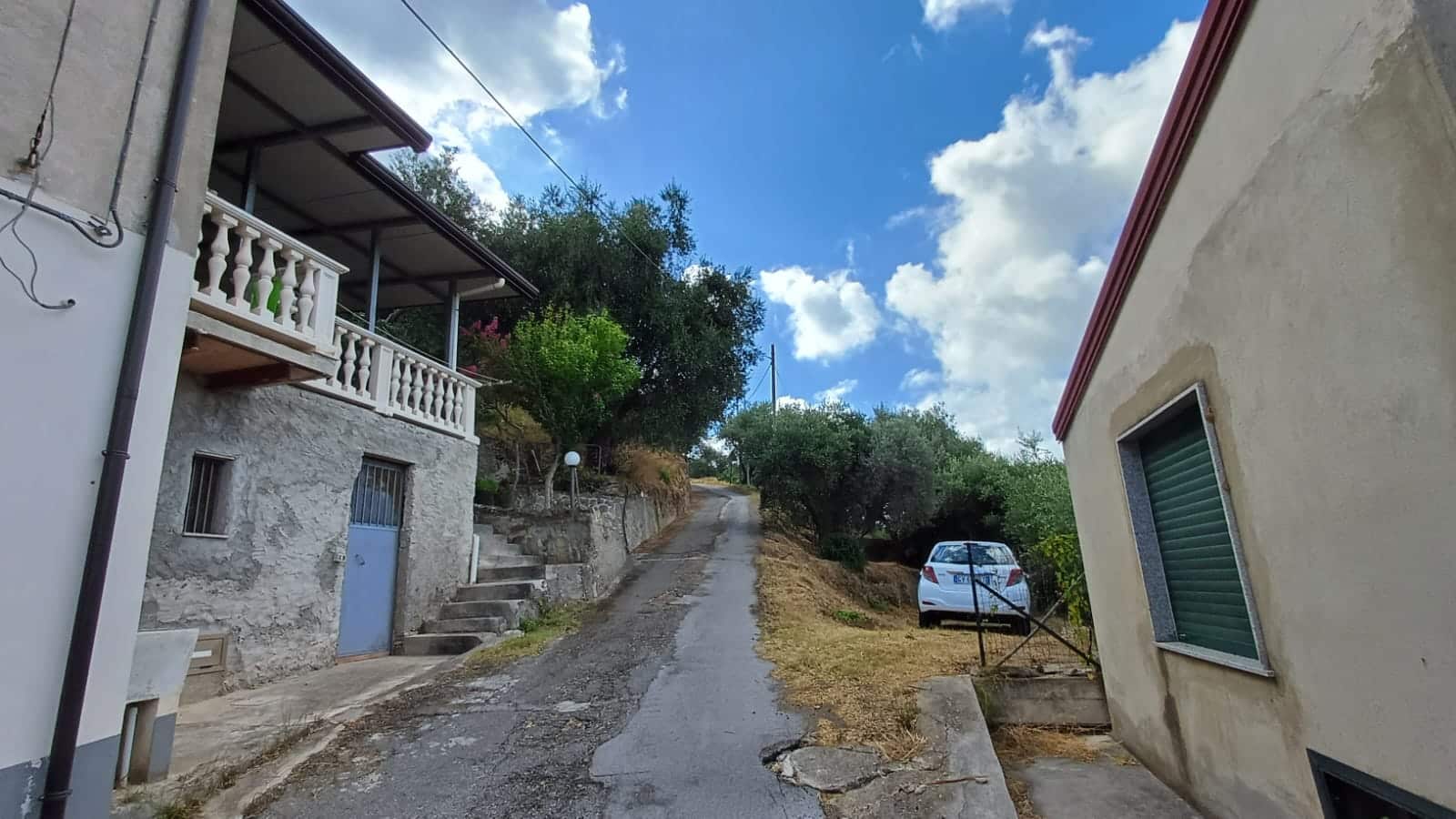 Vecchia strada di montagna con case tradizionali e piante verdi in un paesaggio tranquillo, cielo azzurro con nuvole bianche, ideale per immobiliare e vendita case in collina, NextCasa.