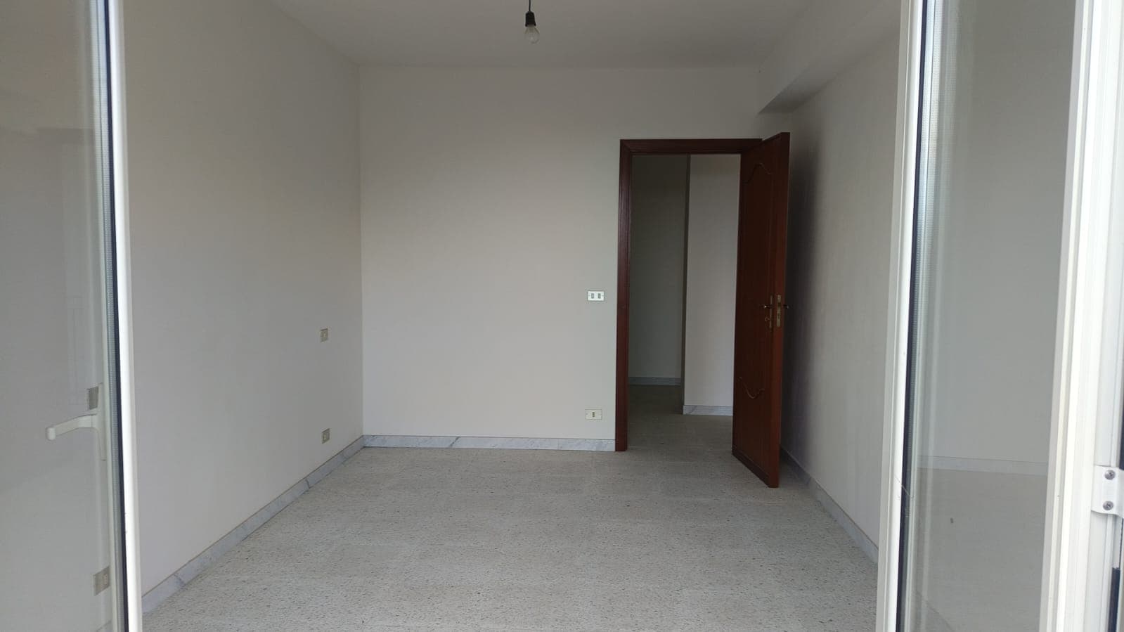 Ampia stanza vuota con parquet chiaro e pareti bianche, porta in legno massiccio e accesso a balcone o terrazza, ideale per immobili di nuova costruzione o ristrutturati, luminoso e funzionale.