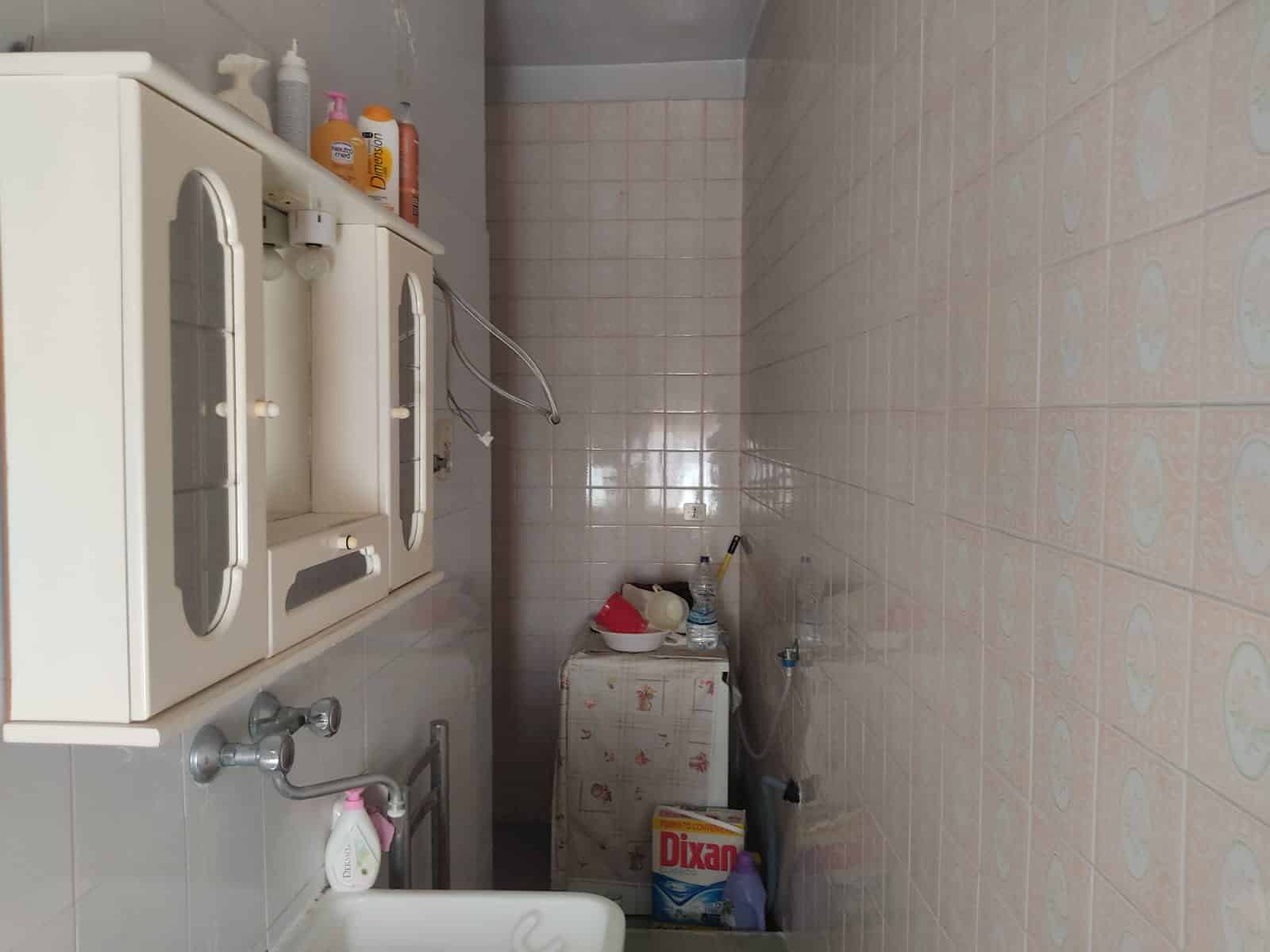 Lavanderia piccola con armadietto bianco, detersivi e prodotti per la pulizia, pavimenti in piastrelle chiare, servizi utili per affitto o vendita, ideale per appartamenti o case.