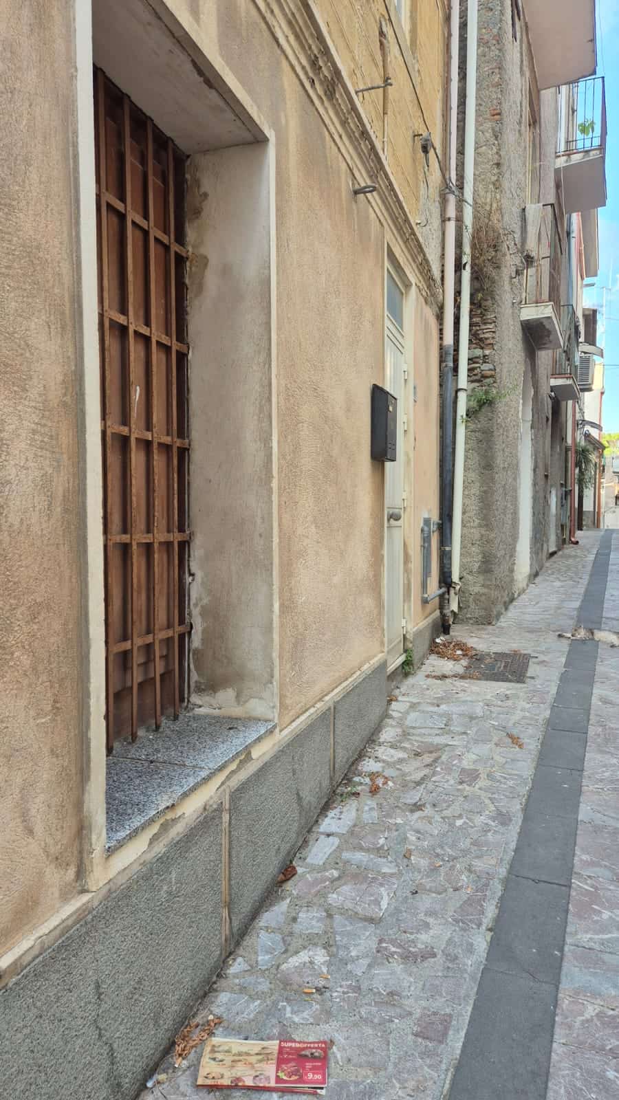 Finestra senza vetri con sbarre di ferro su parete in stili variable e facciata in pietra e intonaco, in strada urbana con pavimentazione in ciottoli e mattoni, contesto di quartiere storico.