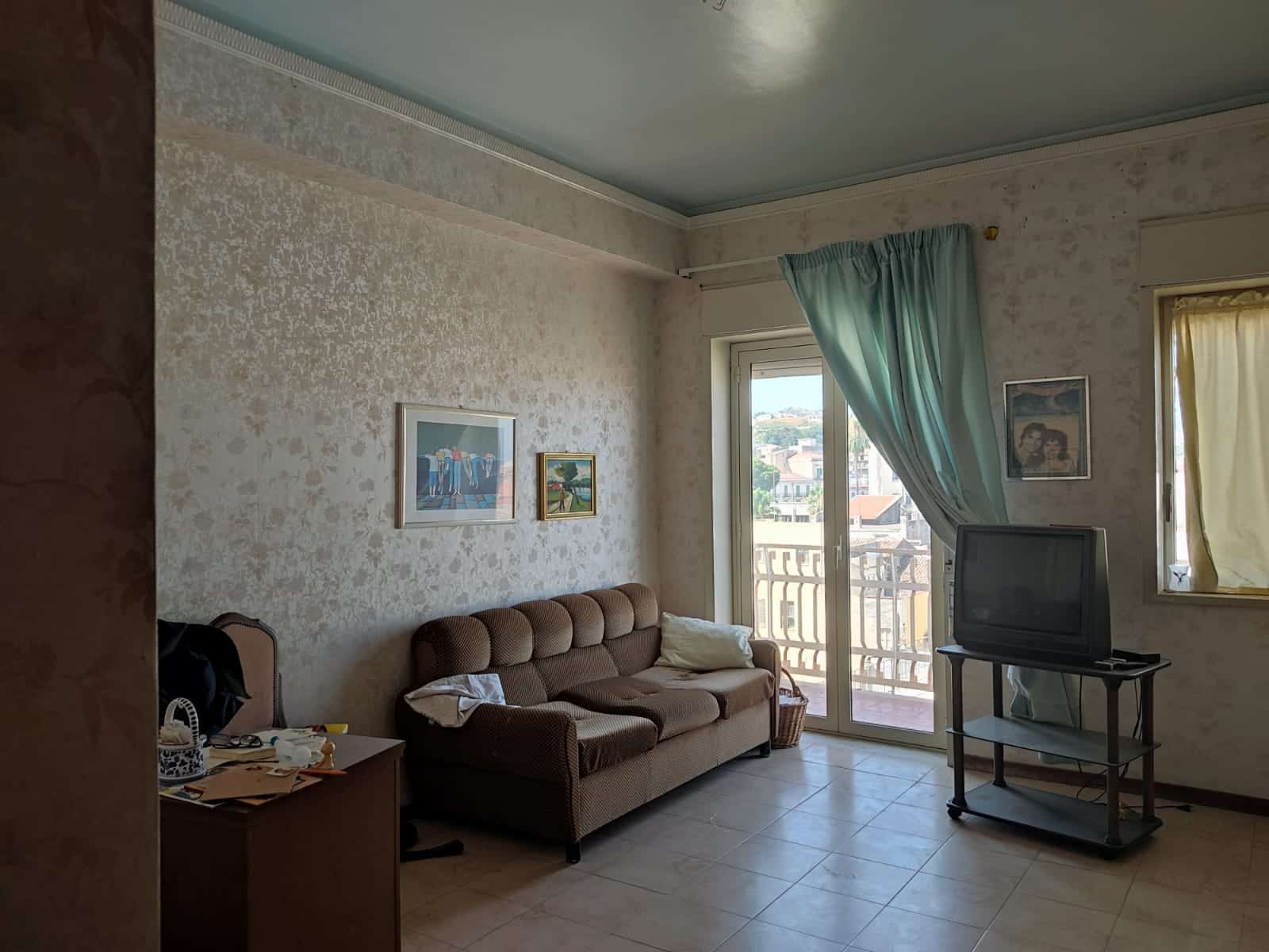 Soggiorno con divano, televisore e balcone con vista, arredamento semplice, pareti decorate, luce naturale, ambiente domestico accogliente e luminoso.