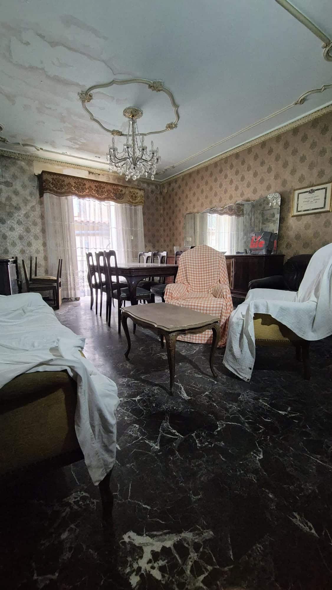 Sofa e poltrone in stile classico con copridivano, sala da pranzo elegante con tavolo e sedie in legno, arredamento vintage con specchio grande e lampadario a cristalli, pavimento in marmo scuro, ambiente raffinato e accogliente.
