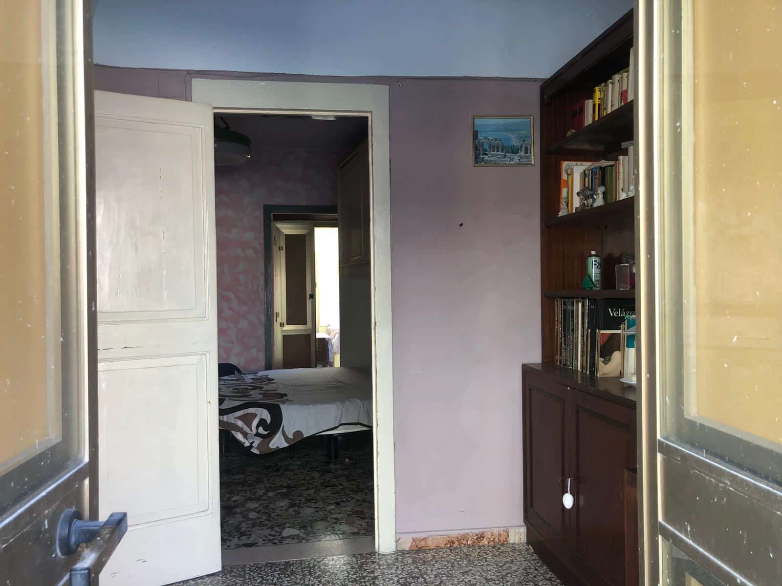 Stanza con libreria in legno e porta d'ingresso, vista su camera da letto con letto e decorazioni, pavimento in piastrelle, stile classico, ambiente abitativo accogliente e pratico.