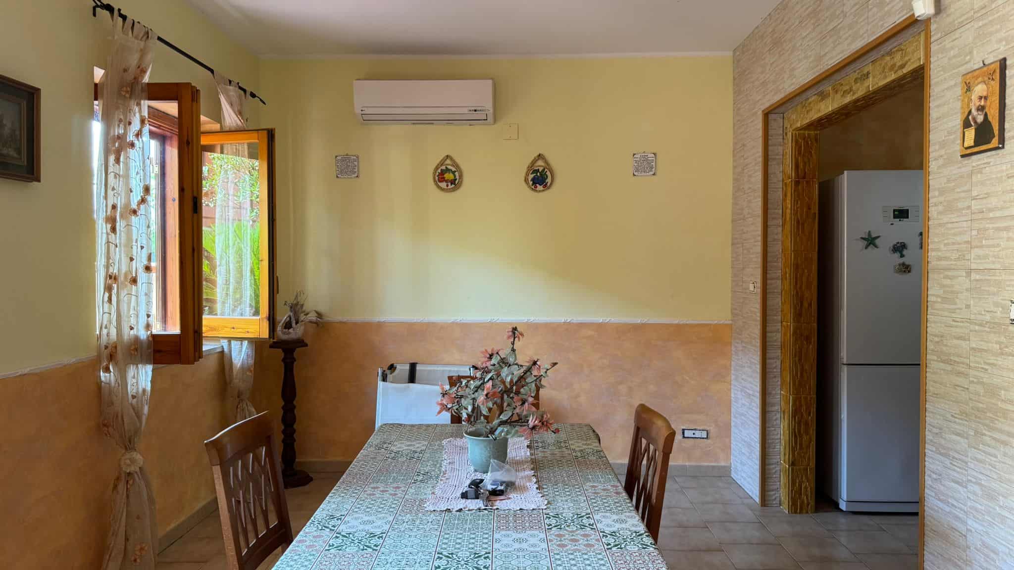 Finestra aperta nella cucina con tende di pizzo, pareti colorate, tavolo da pranzo con fiori secchi in vaso, cucina moderna con frigorifero in vista, ambiente luminoso e accogliente.
