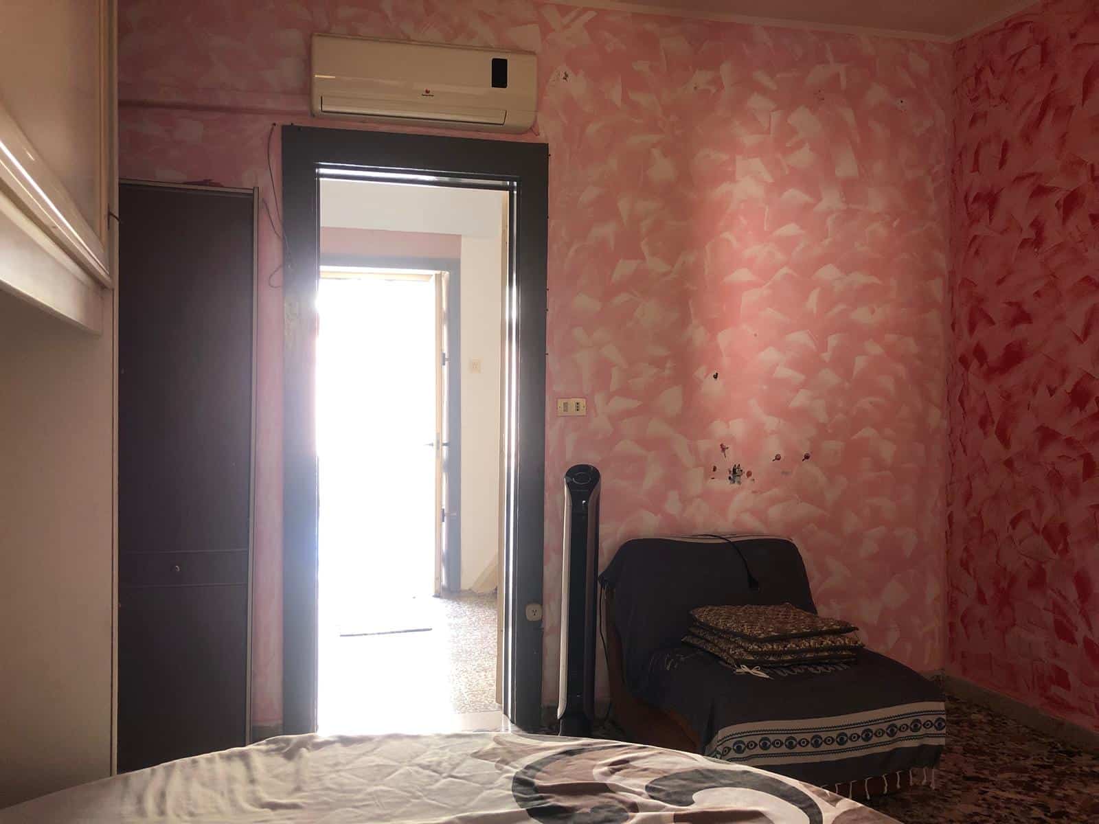Condizione dell'immagine di un interno di casa con pareti rosa, compresa una porta di accesso a una stanza luminosa, aria condizionata, ventilatore, divano e arredi di base, utilizzata per annunci immobiliari NextCasa.