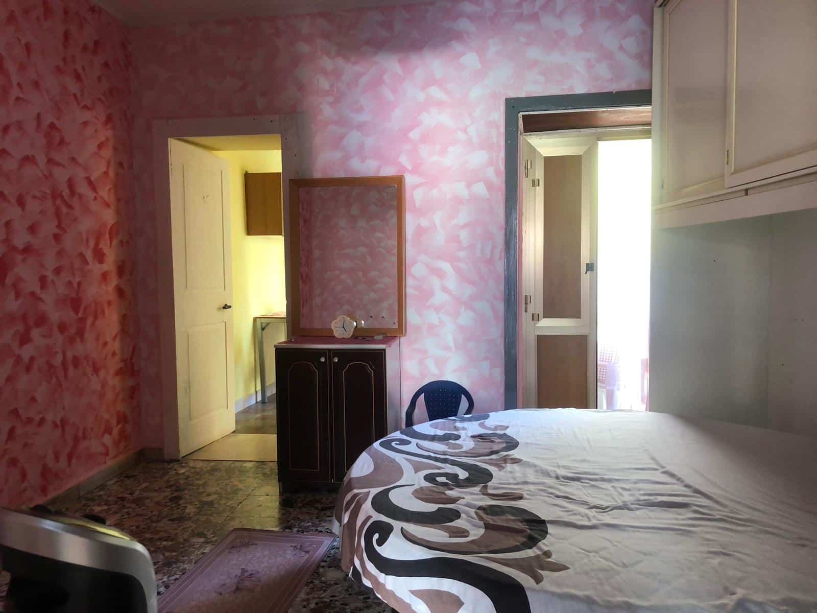 Stanza da letto con pareti dipinte in rosa, letto con copriletto decorativo, finestra con porta di accesso a un balcone o terrazza, arredo classico e atmosfera accogliente.