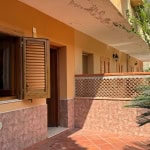 Finestra con persiane in legno su un appartamento con balcone decorato con piastrelle rosse, circondato da piante verdi, in un ambiente luminoso e soleggiato.