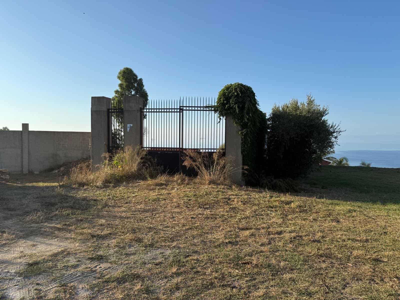 Cancello in ferro battuto con colonne in cemento e vegetazione lussureggiante, vista panoramica sul mare, accesso esterno a proprietà residenziale o terreno rurale, atmosfera tranquilla e naturale, NextCasa.