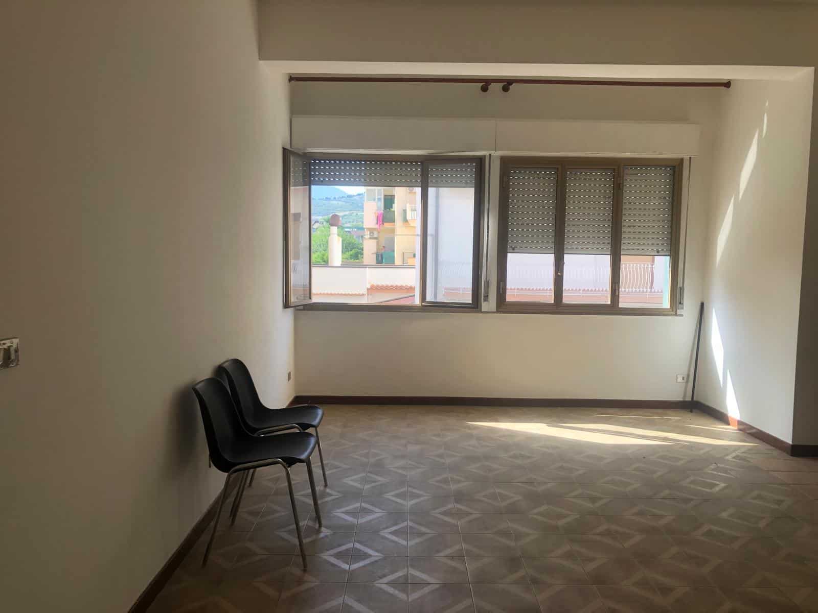 Finestra con persiane in appartamento, spazio abitativo vuoto, luminoso, con sedie nere, pavimento in piastrelle geometriche, vista su edificio e natura, ideale per appartamenti in vendita o affitto, NextCasa.