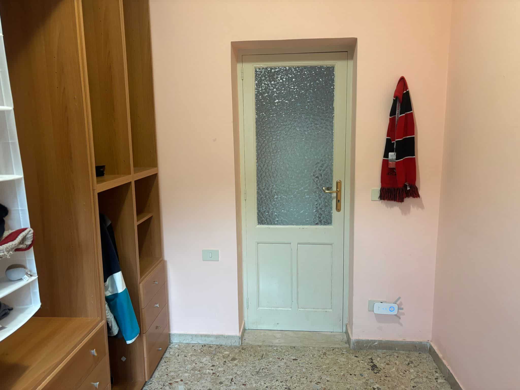 Porta in legno bianca con vetro smerigliato, armadio in legno con ripiani e cassetti, sciarpa rossa e nera appesa alla parete, muro dipinto di rosa chiaro, pavimento in marmo beige, presa elettrica e interruttore.