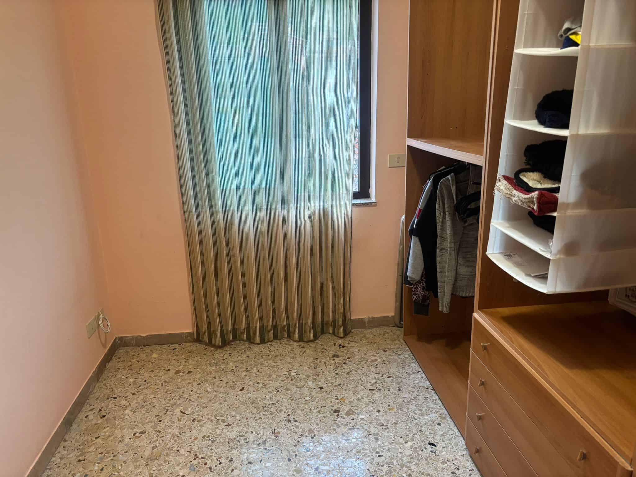 Armadio a muro con abiti e scaffali in legno in camera da letto. Pareti rosa, finestra con tende a righe e pavimento in parquet. Soluzione di arredamento per camere funzionali e ordinate.