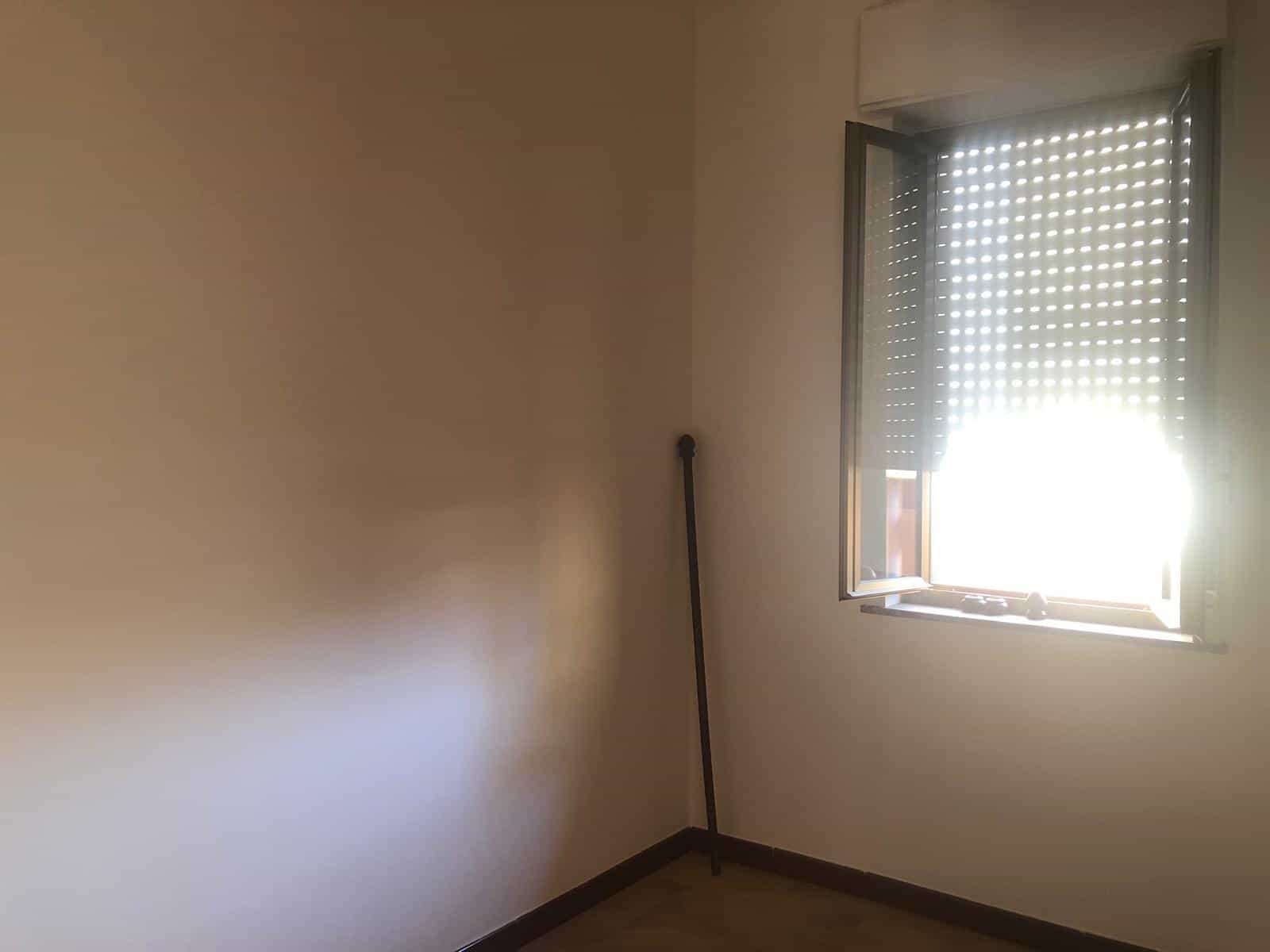 Finestra con tapparella in una stanza vuota di un appartamento, luce naturale entra dalla finestra, pareti beige e pavimento in legno chiaro, ambiente luminoso e minimale.