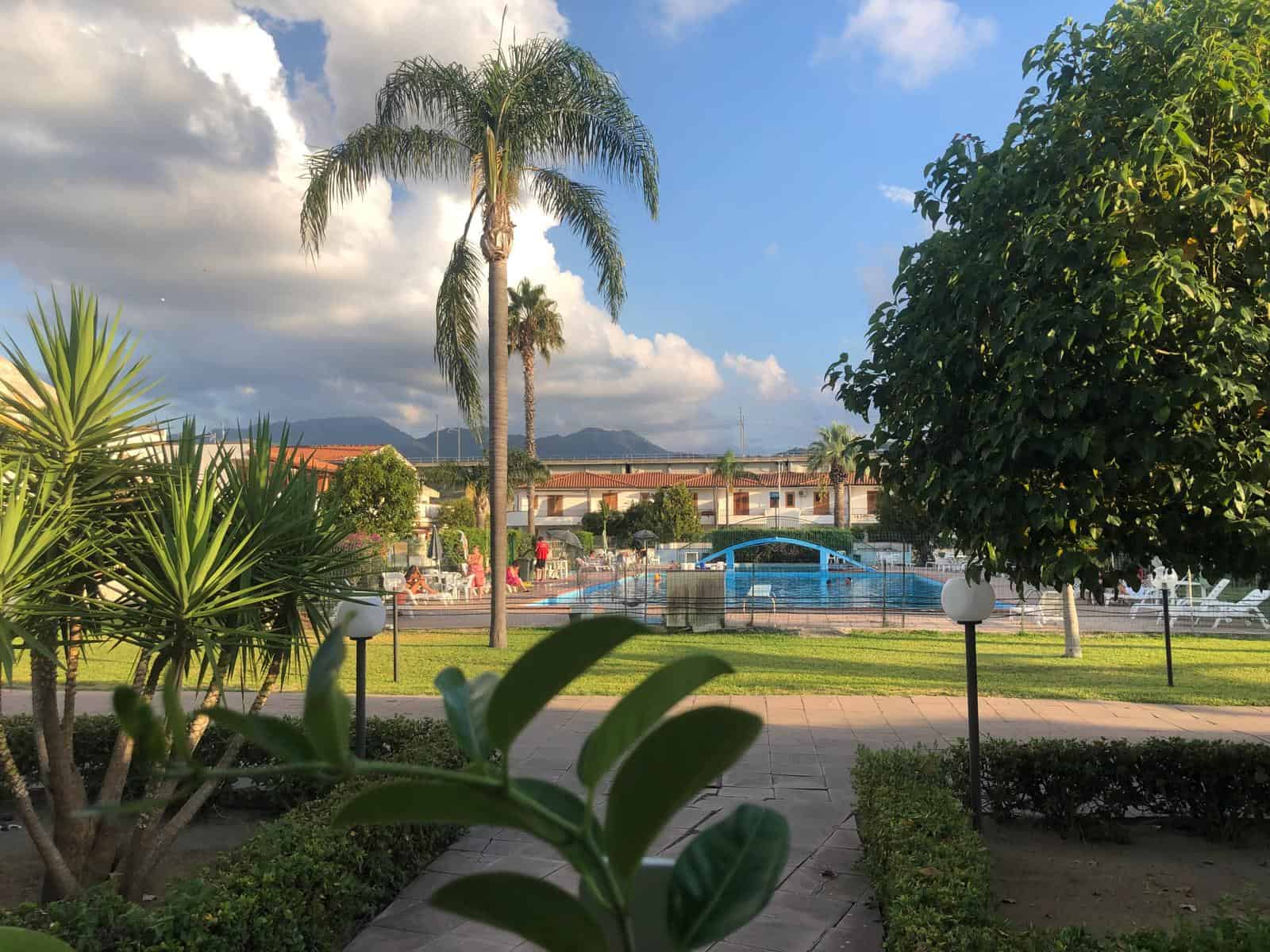 Piscina esterna in un complesso residenziale con piante tropicali, palme e giardino ben curato, panorama montano sullo sfondo, atmosfera rilassante e soleggiata, ideale per vacanze e relax.