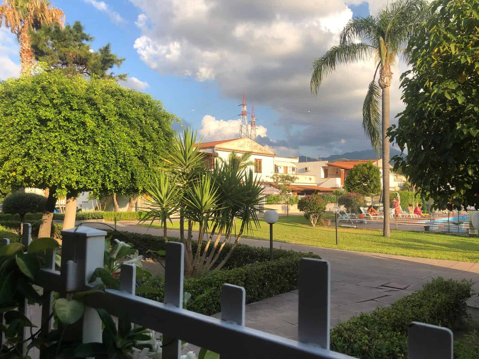 Verde pubblico e giardino condominiale con palme, arbusti e panchine sotto un cielo nuvoloso, ambientazione di un residence prossimo alla spiaggia o piscina, immagini di ambienti esterni di case NextCasa.