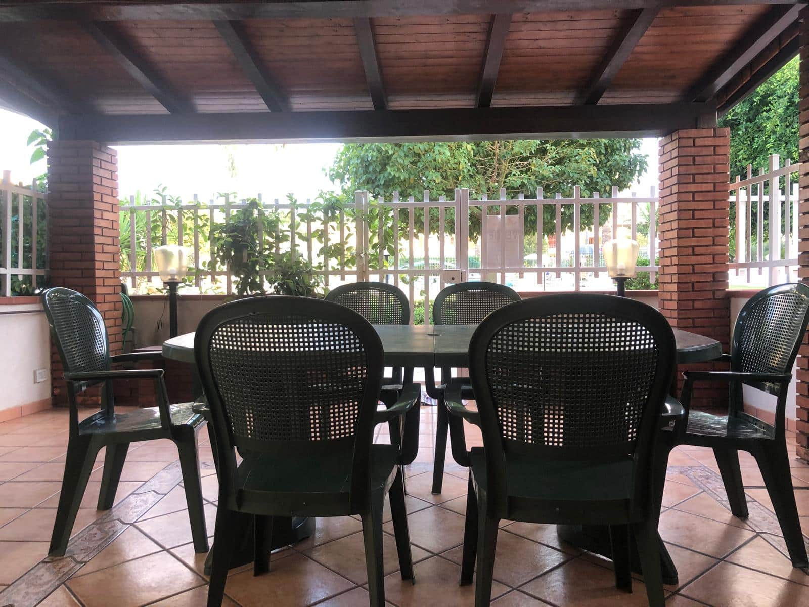 Tavolo da pranzo all'aperto con sedie in plastica sotto una tettoia in legno, contornato da muretti in mattoni rossi e recinzione bianca, in un'area esterna luminosa e verdeggiante.