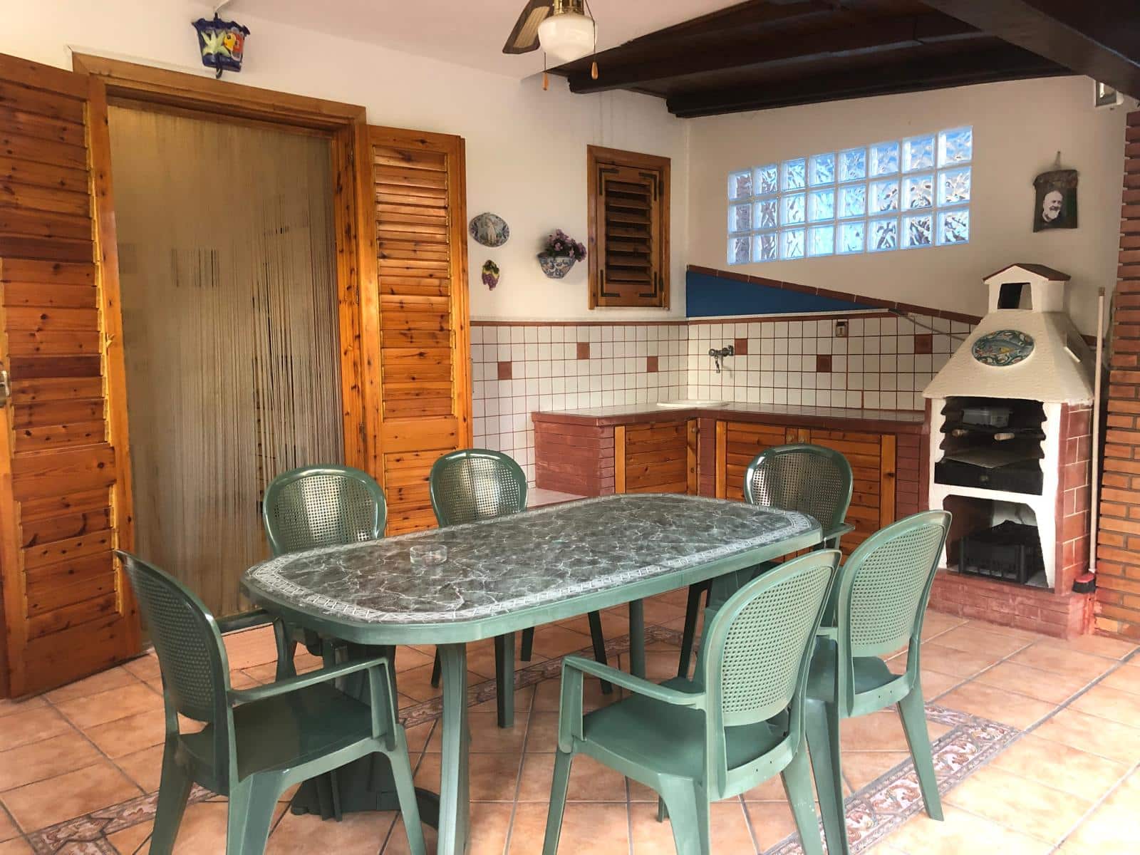 Mensa da pranzo in stile rustico con tavolo in marmo, sedie in plastica verde, camino in mattoni e cucina con piastrelle bianche e dettagli in legno. Ambiente accogliente e rustico.