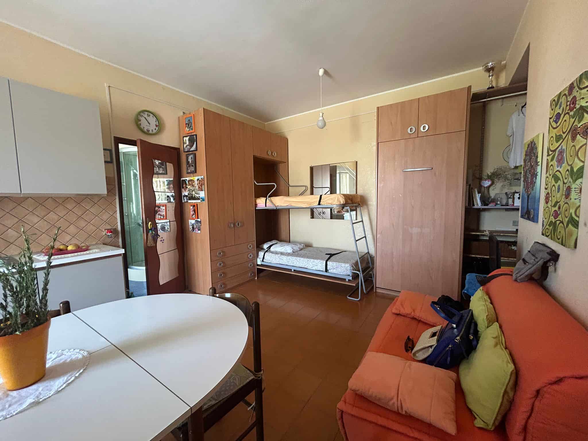 Camera da letto e soggiorno in appartamento con mobili in legno, letto a castello, divano arancione, tavolo bianco, decorazioni colorate, orologio digitale, spazio funzionale e accogliente.