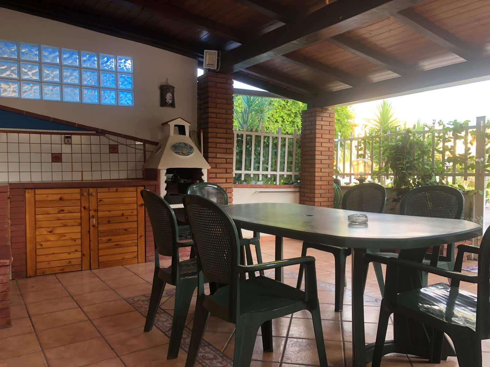 Accogliente patio esterno con zona barbecue, tavolo da pranzo, sedie in plastica, mattonelle in ceramica, tetto in legno, decorazioni in mattoncini rossi, circondato da piante verdi e recinzione metallica, ideale per relax e convivialità.