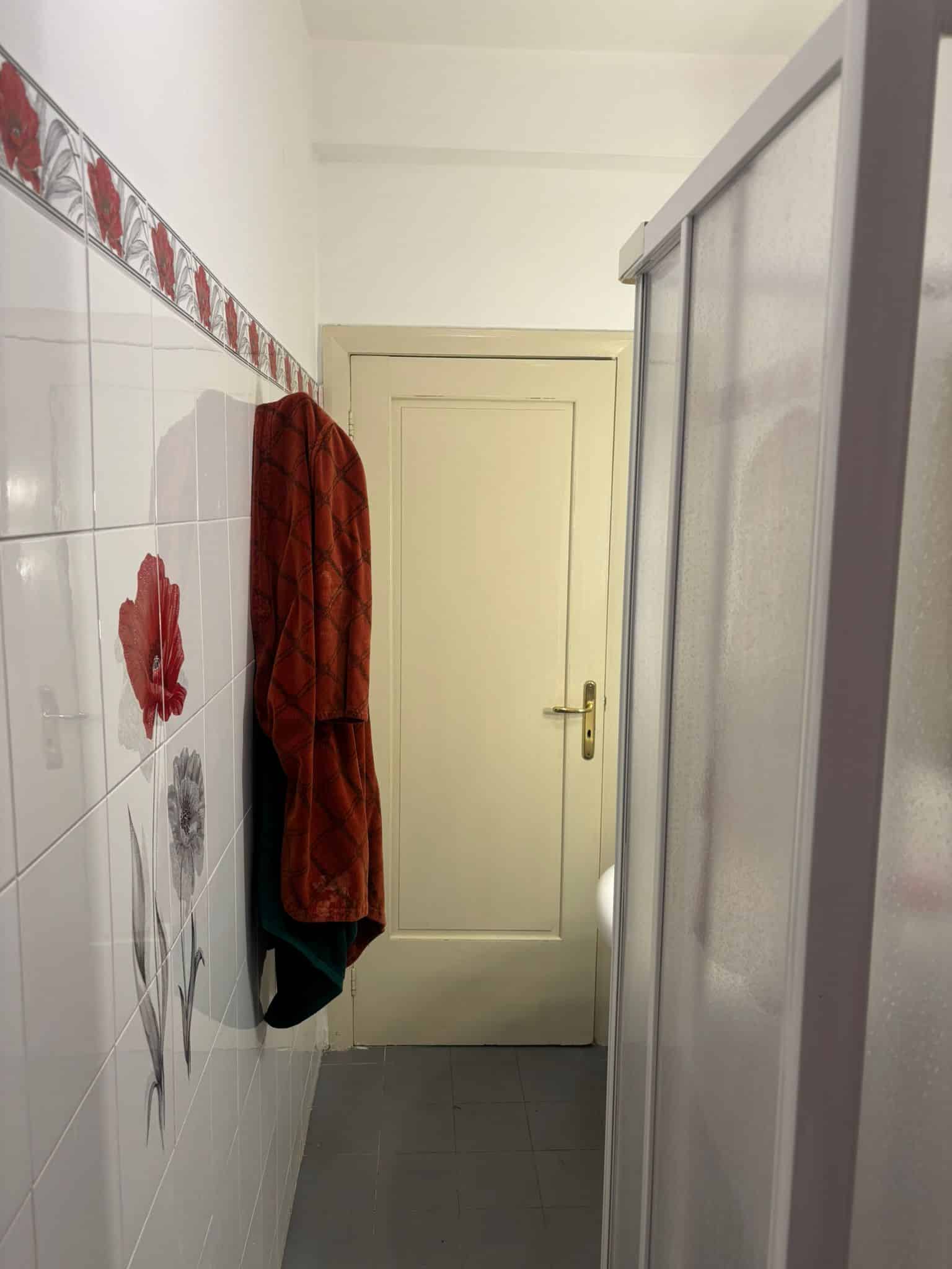Panni appesi in un bagno con pareti rivestite di piastrelle bianche con decori floreali rossi e grigi, porta in elettrodomestico e parete chiara.