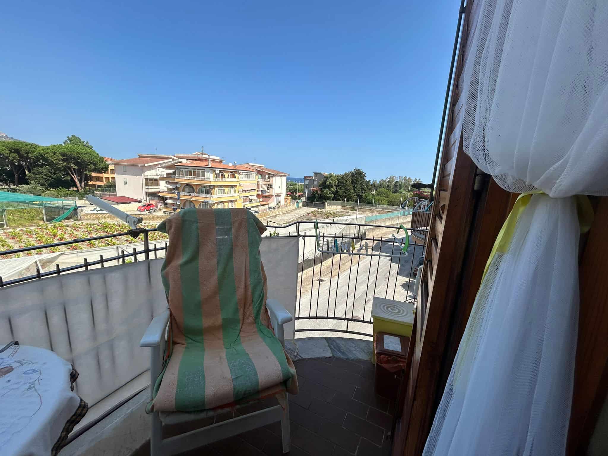 Balcone con vista sul cielo e sui palazzi residenziali, arredato con sedia e tende di tessuto leggero, ideale per relax e godersi il panorama urbano.