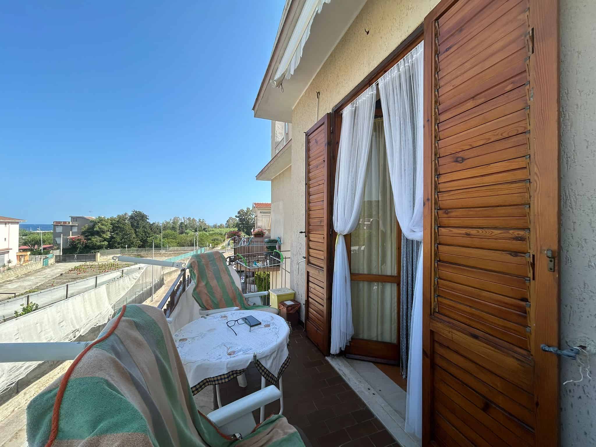 Balcone di un appartamento con soluzione di tende legno e tenda leggera, vista mare e paesaggio con verde e cielo sereno, ideale per case vacanza o residenze nel mediterraneo.