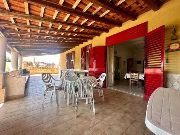 Balcone con vista, arredato con tavoli e sedie, con tettoia in legno, porte e persiane rosse, in stile rustico, ideale per relax e pranzi all'aperto, di fronte a un giardino e un paesaggio rurale.