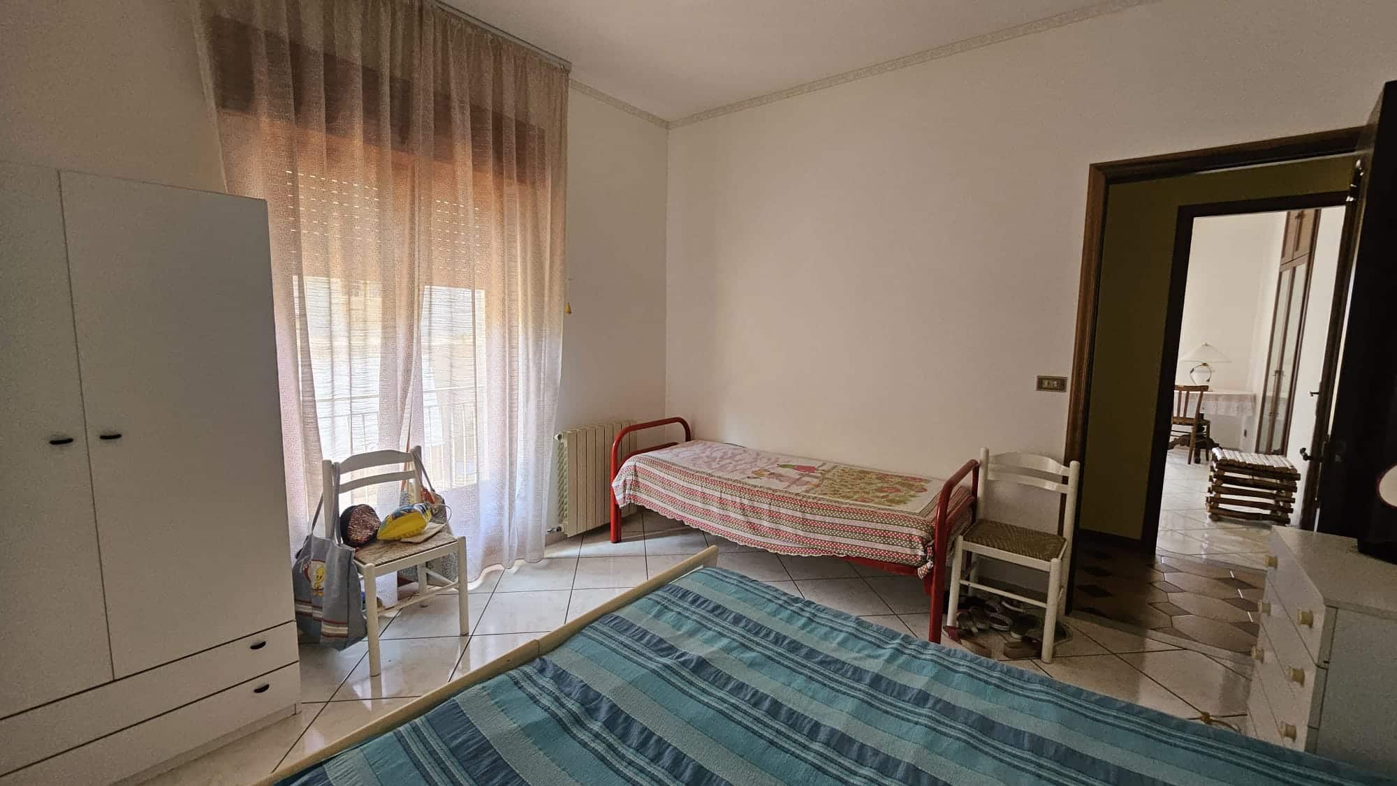 Letto singolo con copriletto a righe blu in camera da letto. Finestra con tende leggere color pesca lascia entrare la luce naturale. Arredamento semplice e pratico in stile classico.