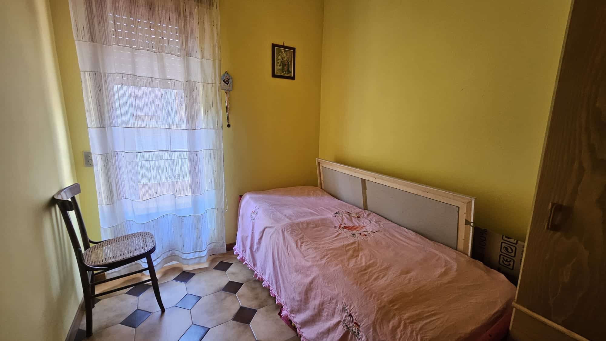Letto singolo in camera con pareti gialle, tende leggere e pavimento in piastrelle beige e marroni, arredamento semplice ed economico, ideale per affitti brevi o studenti.