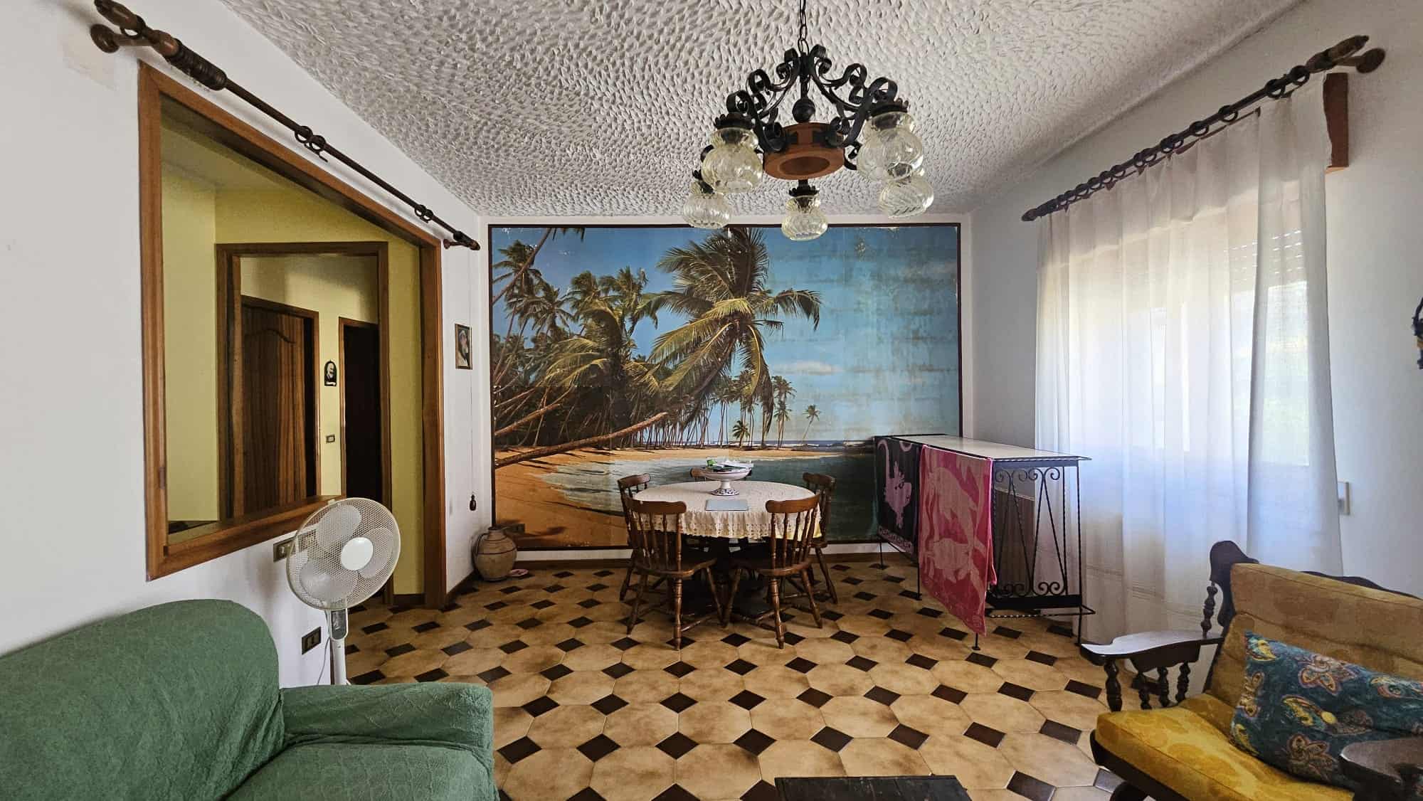 Spazio soggiorno con grande parete decorata con foto di spiaggia tropicale, divano verde, poltrona gialla, tavolo rotondo con sedie in legno, lampadario antico e finestra con tende leggere, atmosfera accogliente e familiare.