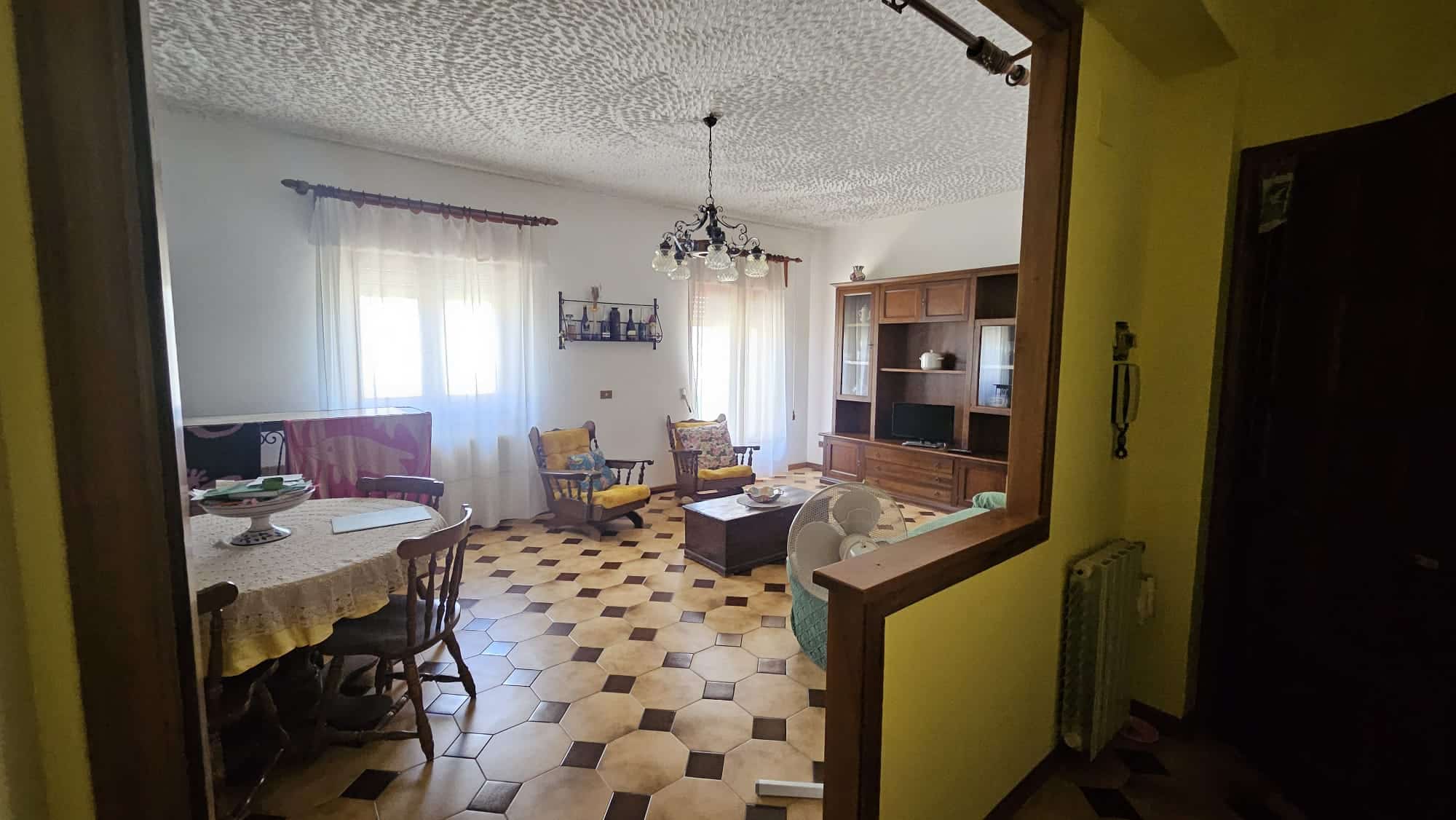 Vista soggiorno con mobili in legno, sedie e divano, grande finestra con tende leggere, pavimento in piastrelle geometriche, atmosfera accogliente e tradizionale.