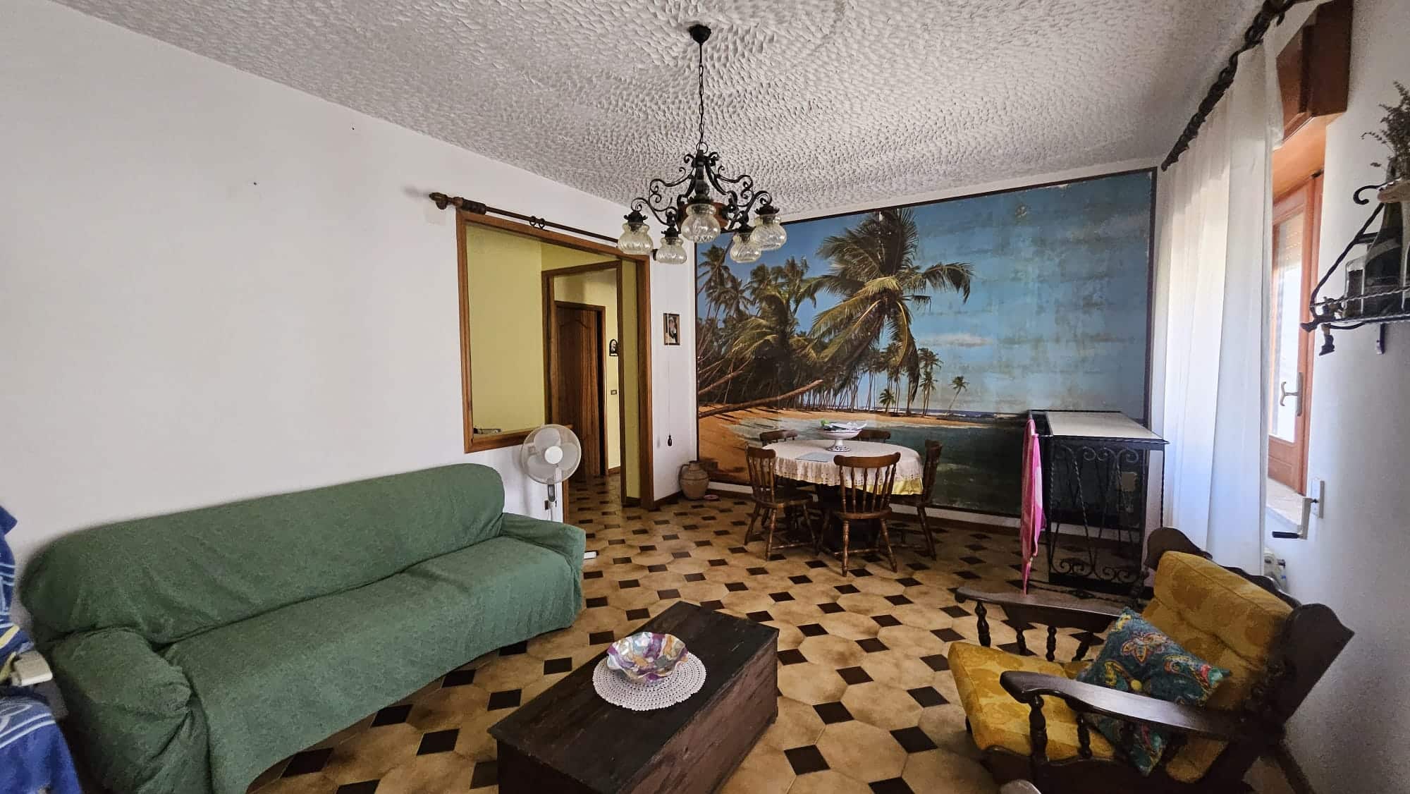 Soggiorno con grandi parete decorata con immagine di spiaggia tropicale, arredamento classico, divano verde e poltrona gialla, pavimento in parquet, atmosfera accogliente e panoramica turistica.