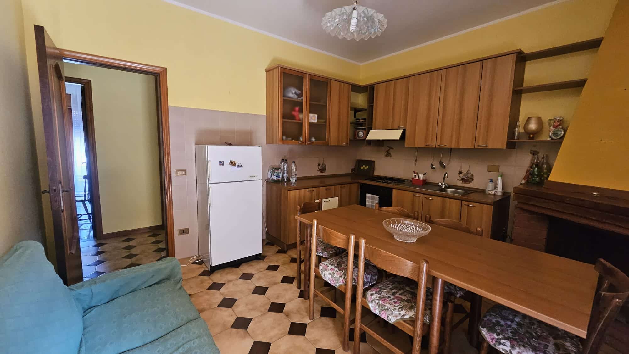 Cucina abitabile con mobili in legno, frigorifero bianco, tavolo in legno con sedie fiori, pareti gialle e beige, arredamento classico, ambiente accogliente, dettagli di cucina e soggiorno, casa in stile rustico.