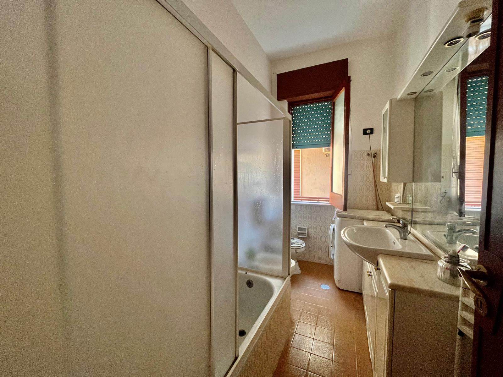 Finestra nel bagno di una casa, con arredo di mobili e lavabo, piastrelle beige e pareti con stile retrò, illuminato naturalmente da una finestra con tapparella verde, in un appartamento in affitto.
