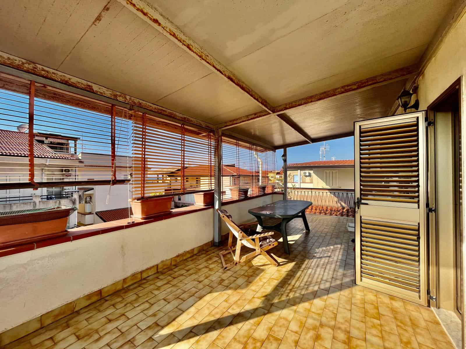 Ampio terrazzo coperto in zona residenziale, ideale per relax e aperitivi, con vista su altre abitazioni, pavimentazione in piastrelle, inferriate e serranda in legno.