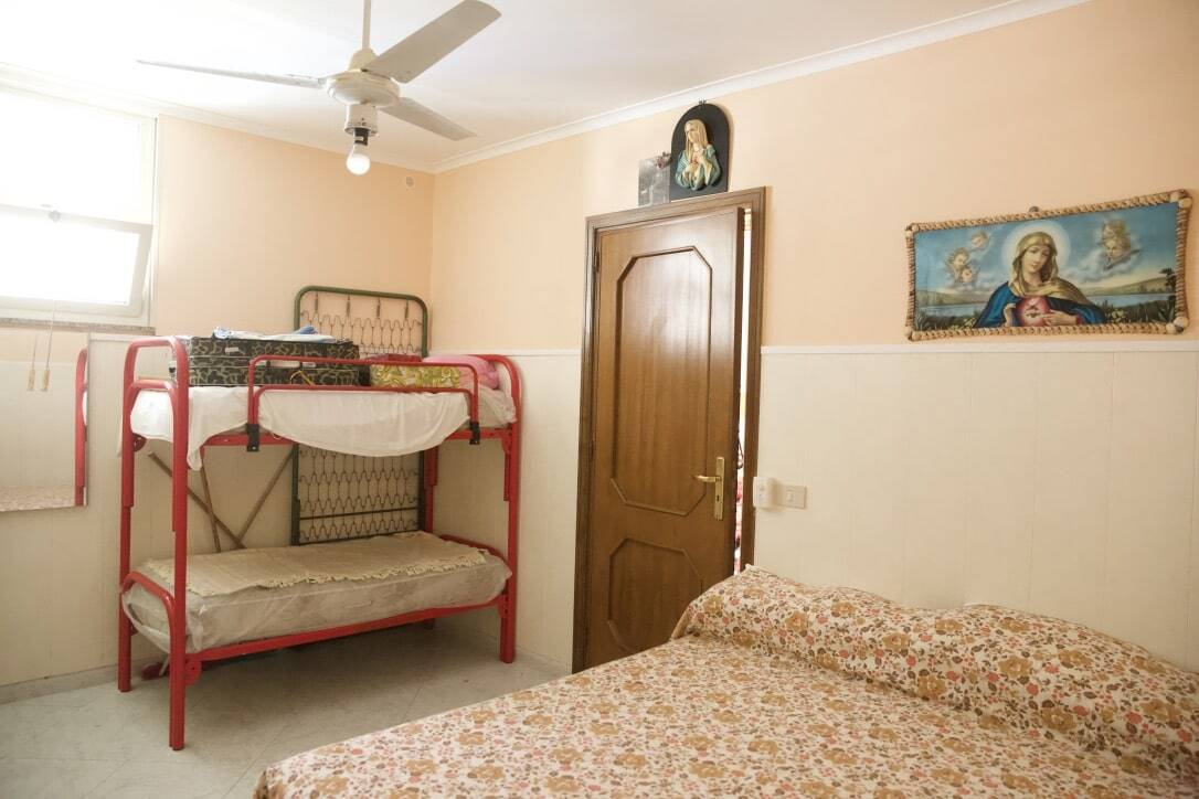Red bunk bed in a cozy bedroom with religious art, ceiling fan, and floral bedspread, perfect for family or student accommodation.