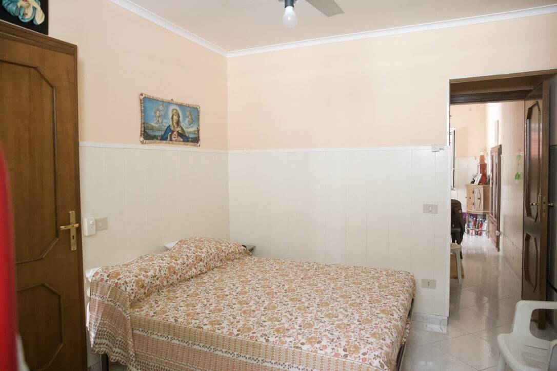 Camera da letto con pareti beige e bianche, arredamento semplice, quadro religioso sopra il letto, ambiente luminoso e accogliente.