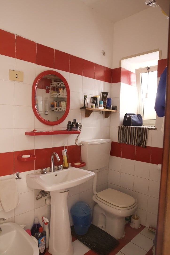 Lavabo bagno con specchio rosso, arredamento di spray, spazzole, prodotti per la cura personale e scaffalature, illuminato da finestra con tende a righe.