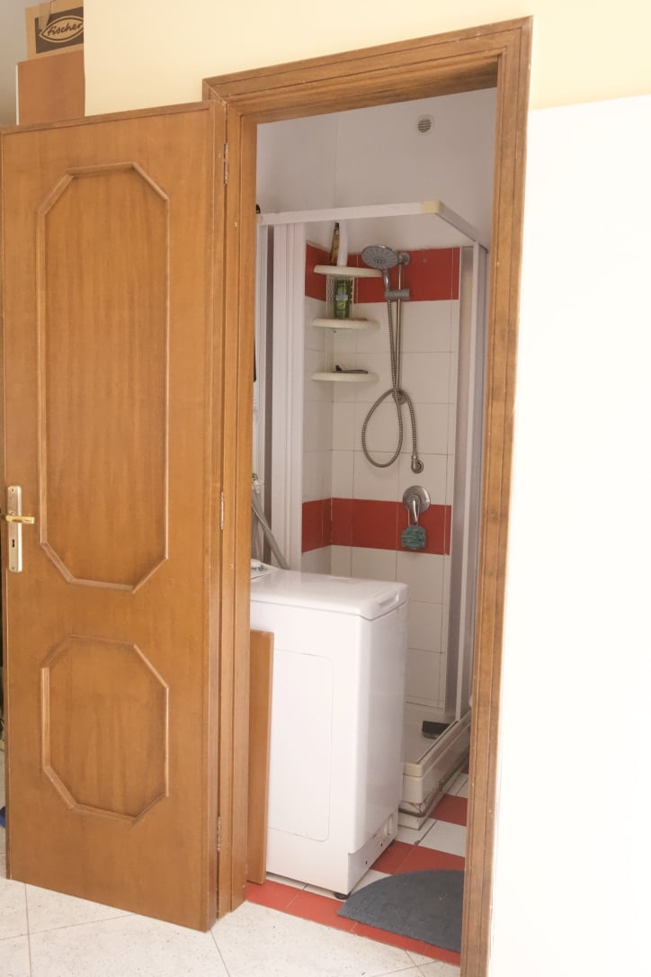 Lavatrice e doccia in un ripostiglio bagno con pareti a righe di azzurro e rosso, accessibile tramite porta in legno. Soluzione funzionale e compatta per spazi interni.