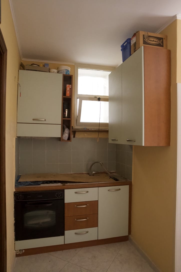 Vecchia cucina compatta con armadietti bianchi e legno, forno elettrico, lavabo sottopensile, finestra piccola naturale, ripiani a muro e scatole di stoccaggio, ambiente di casa funzionale e ordinato.