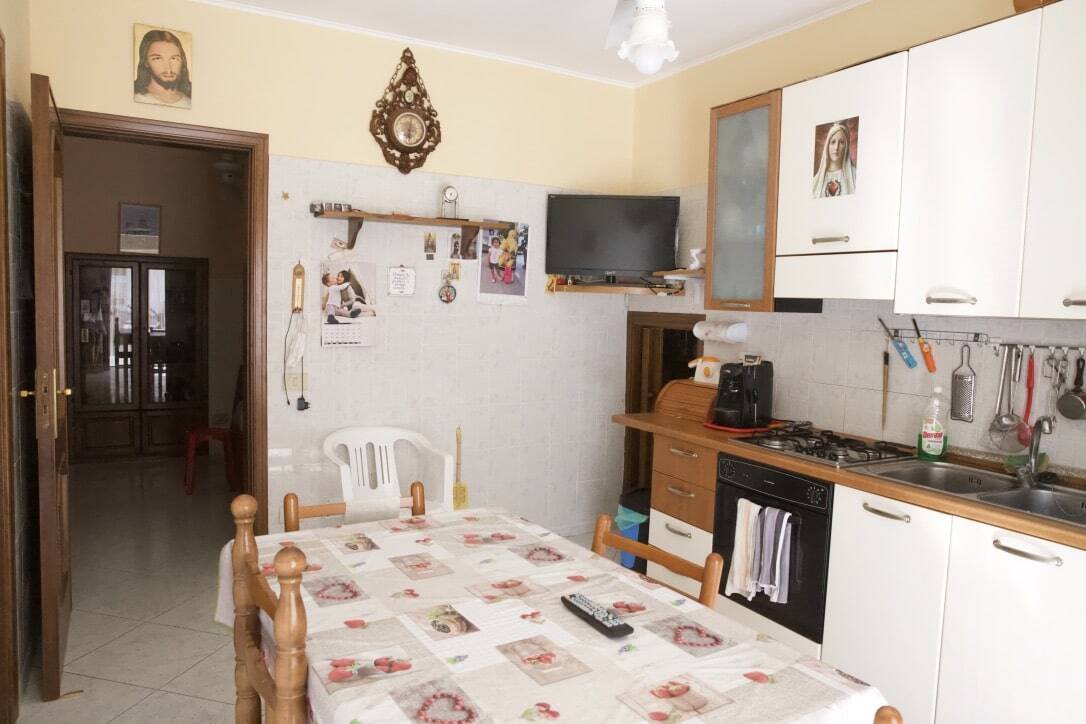 Casalinga cucina abitabile con tavolo, elettrodomestici, mobili in legno e decorazioni religiose, ambiente conviviale e funzionale, ideale per la vita quotidiana e momenti di convivialità domestica.