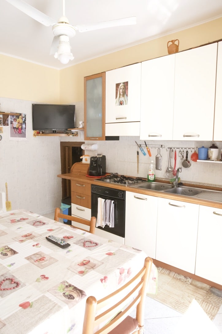 Cucina abitabile con mobili bianchi e accessori cucina, tavolo da pranzo con tovagliato decorato, elettrodomestici, TV e dettagli decorativi, ambiente accogliente e funzionale, ideale per_case prossime alla vendita o affitto, NextCasa.
