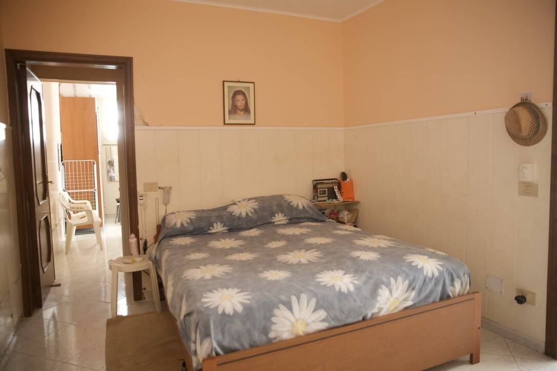 Letto matrimoniale con copriletto a fiori, arredamento semplice, camera da letto luminosa, pareti color pesca e bianche, quadri e accessori vari, ideale per appartamenti di vendita o affitto.